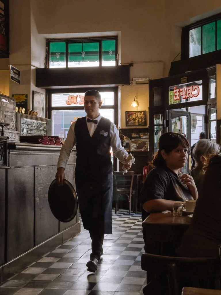 A waiter in a bow tie and apron walking through the vintage interior of Bar Plaza Dorrego in San Telmo, Buenos Aires, with black and white tiled floors, old enamel signs and green stained-glass windows