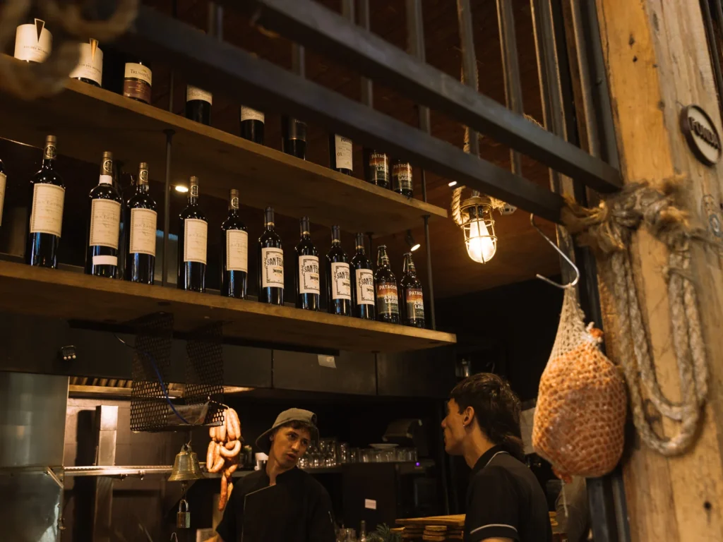 The bar counter of a rustic San Telmo restaurant in Buenos Aires, with shelves of Argentine Malbec wine bottles, a hanging cured meat joint and two staff members talking beside the open kitchen