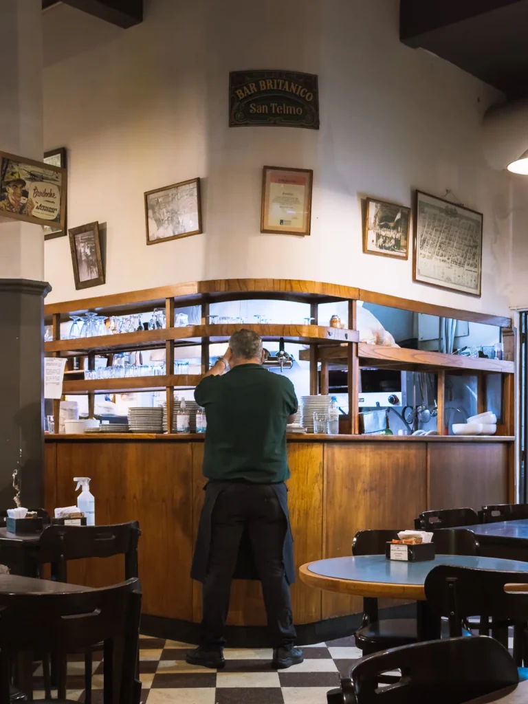 A waiter working behind the wooden bar counter of Bar Británico in San Telmo, Buenos Aires, with a checkered floor, framed vintage photographs on the walls and a sign reading "Bar Británico San Telmo" above