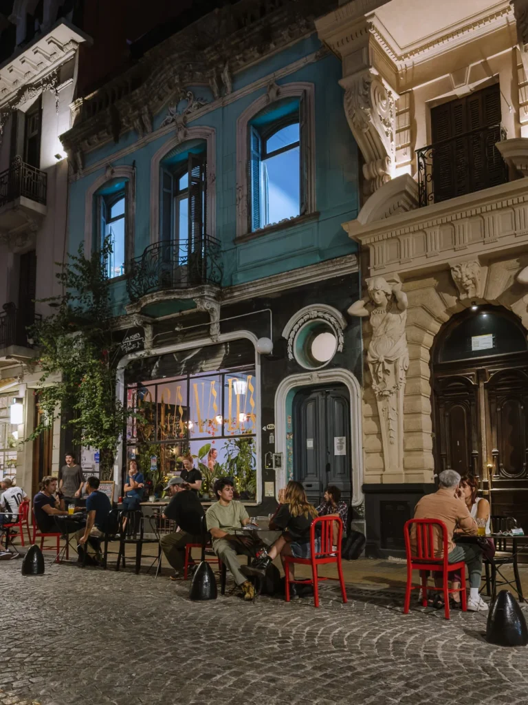 Diners sitting at outdoor tables on red chairs on a cobblestone street in San Telmo, Buenos Aires at night, with a teal Art Nouveau building and ornate Beaux-Arts façade lit up behind them