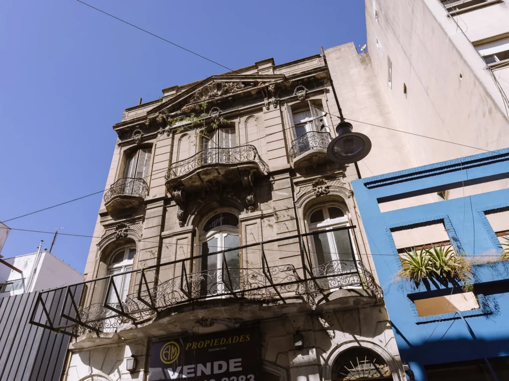 A weathered Beaux-Arts building in San Telmo, Buenos Aires, with ornate stone carvings, wrought-iron balconies and a "Se Vende" (for sale) sign on the ground floor, next to a bright blue shopfront