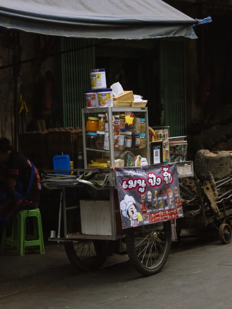 A street food cart loaded with jars and packaged goods parked in a Bangkok back alley.