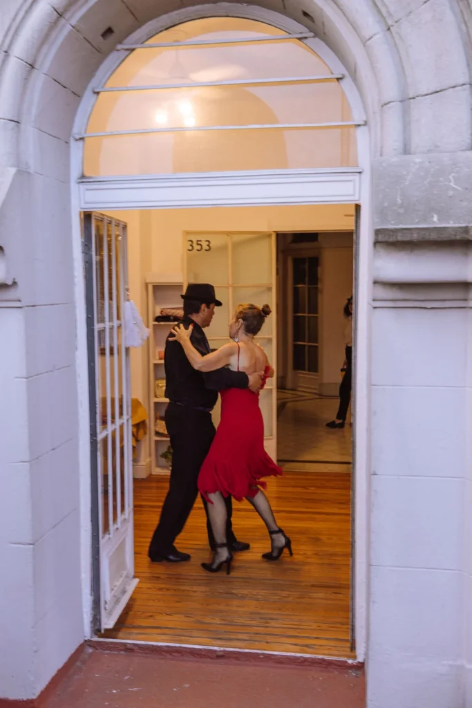 A tango couple dancing in the arched doorway of a building in San Telmo, Buenos Aires, with the woman in a red dress and the man in black leading the dance, watched by a spectator in the background