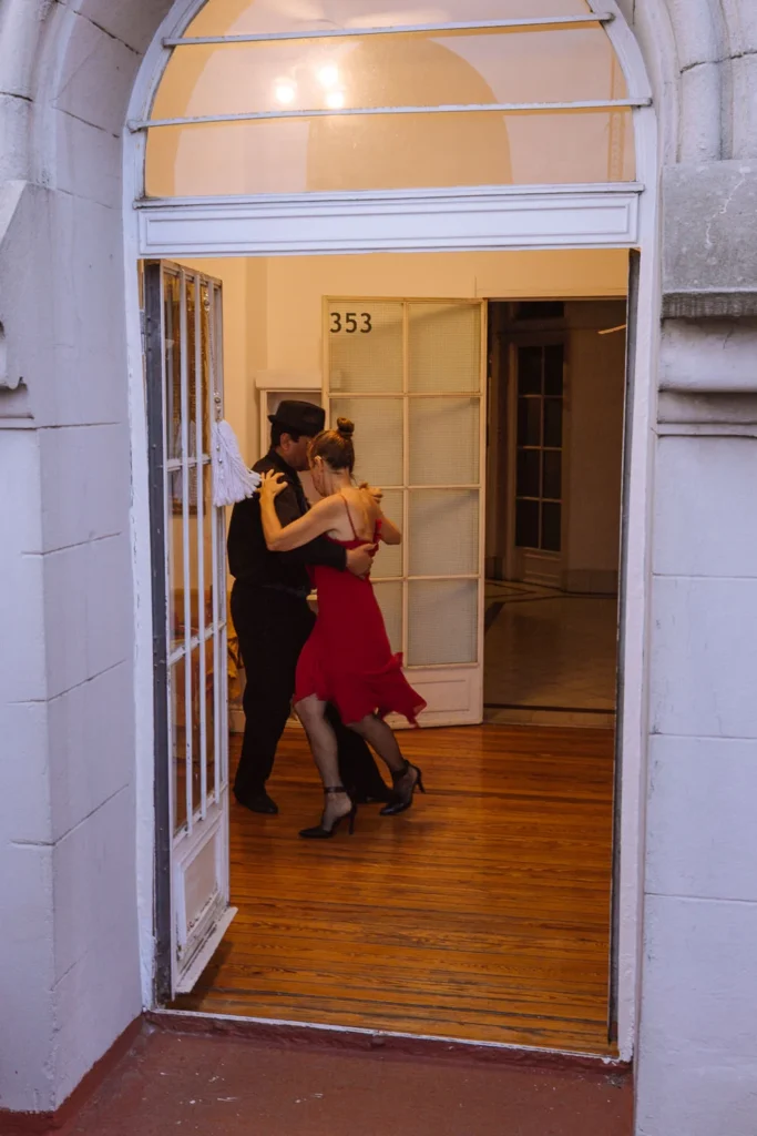 A tango couple dancing inside a doorway in San Telmo, Buenos Aires — the woman in a flowing red dress and heels, the man in black with a fedora, captured mid-movement on a polished wooden floor