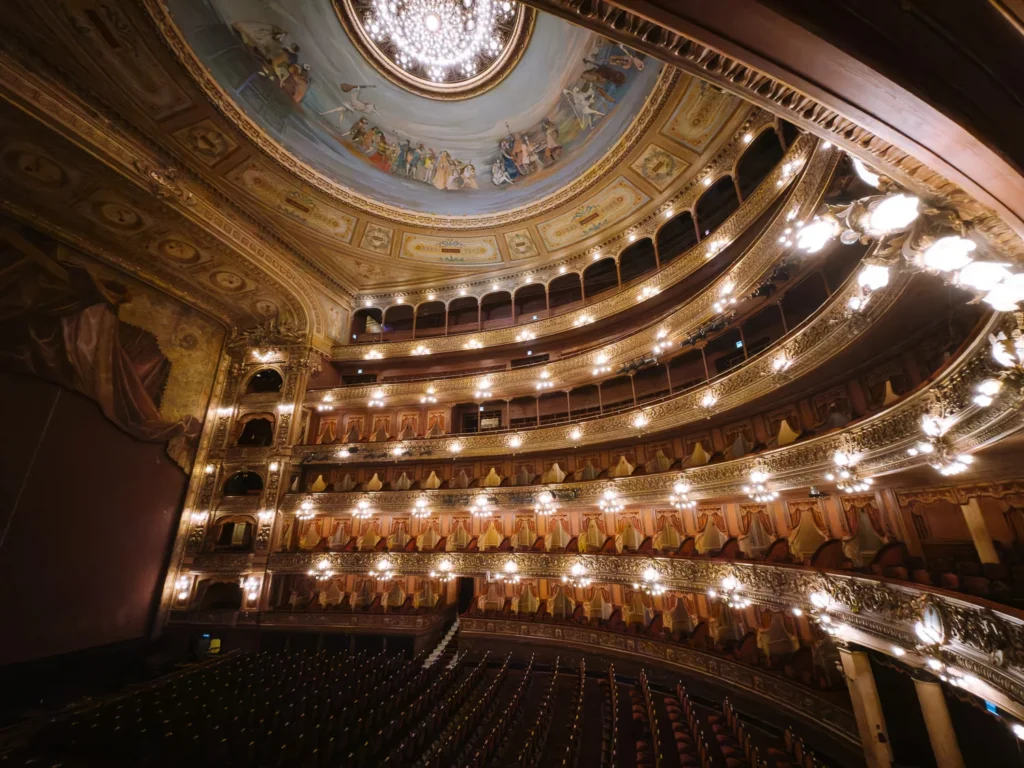 The full interior of the Teatro Colón auditorium in Buenos Aires, showing seven tiers of gilded balconies, the painted dome ceiling and rows of empty stalls seats