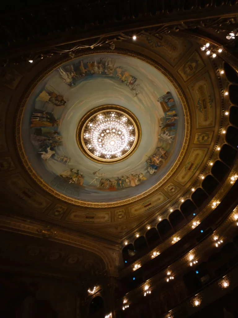 Looking up at the painted dome ceiling and central chandelier of the Teatro Colón in Buenos Aires, surrounded by tiered balconies and ornate gold detailing