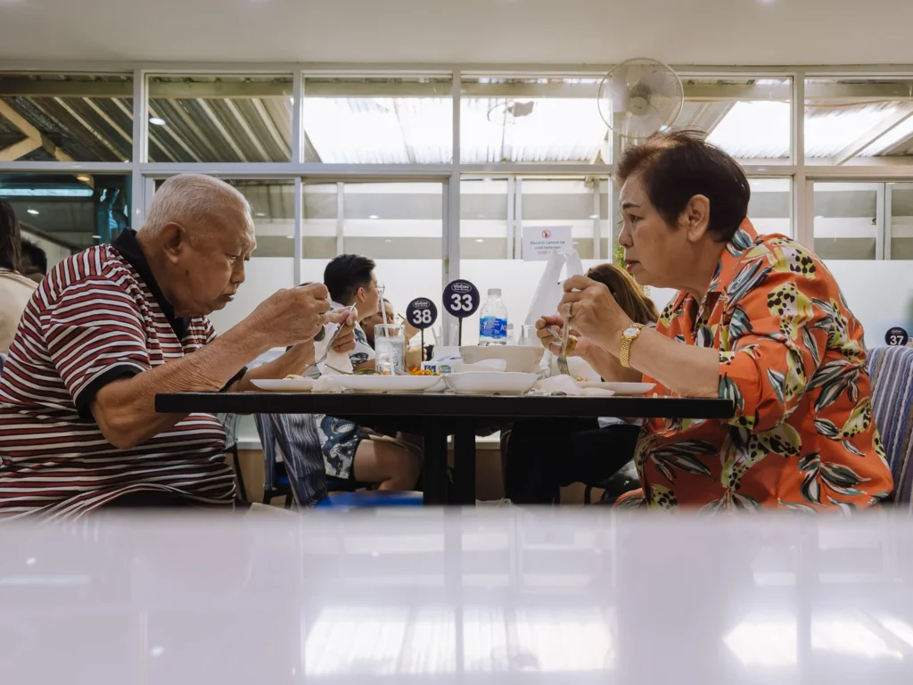 Two people eating together at a busy Bangkok food court, with numbered table markers and other diners visible in the background.