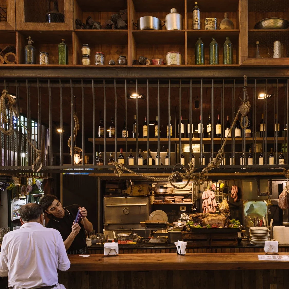 The rustic interior of a Buenos Aires parrilla, with hanging sausages, cuts of meat on display, wine bottles lining the shelves, and diners seated at a wooden bar counter