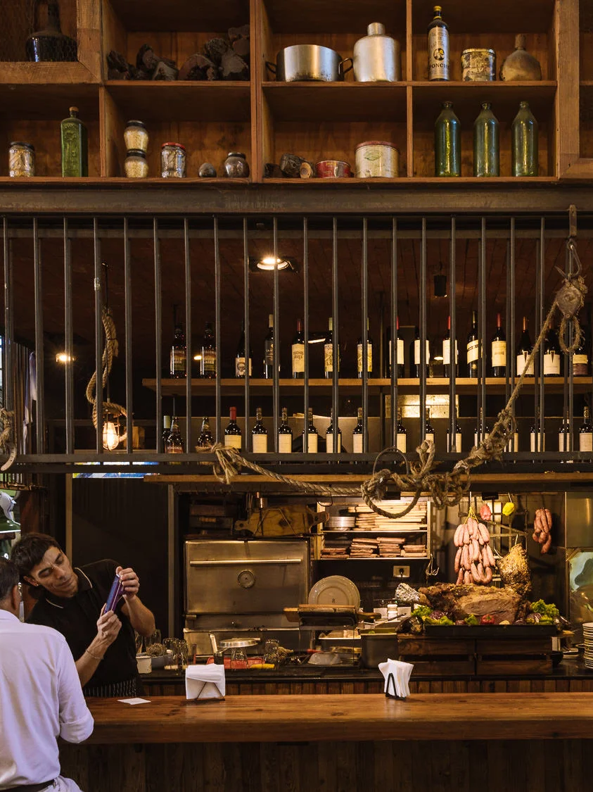 The rustic interior of a Buenos Aires parrilla, with hanging sausages, cuts of meat on display, wine bottles lining the shelves, and diners seated at a wooden bar counter