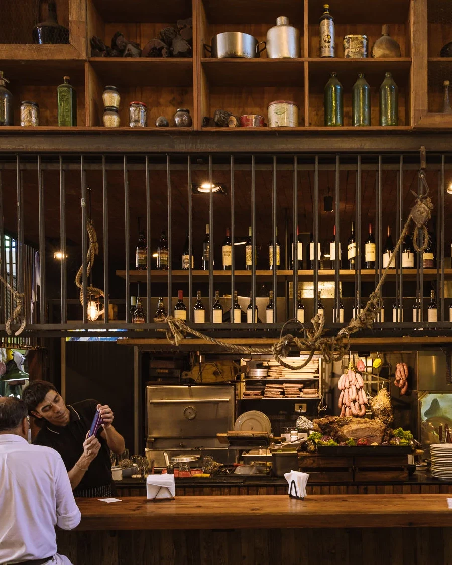 The rustic interior of a Buenos Aires parrilla, with hanging sausages, cuts of meat on display, wine bottles lining the shelves, and diners seated at a wooden bar counter