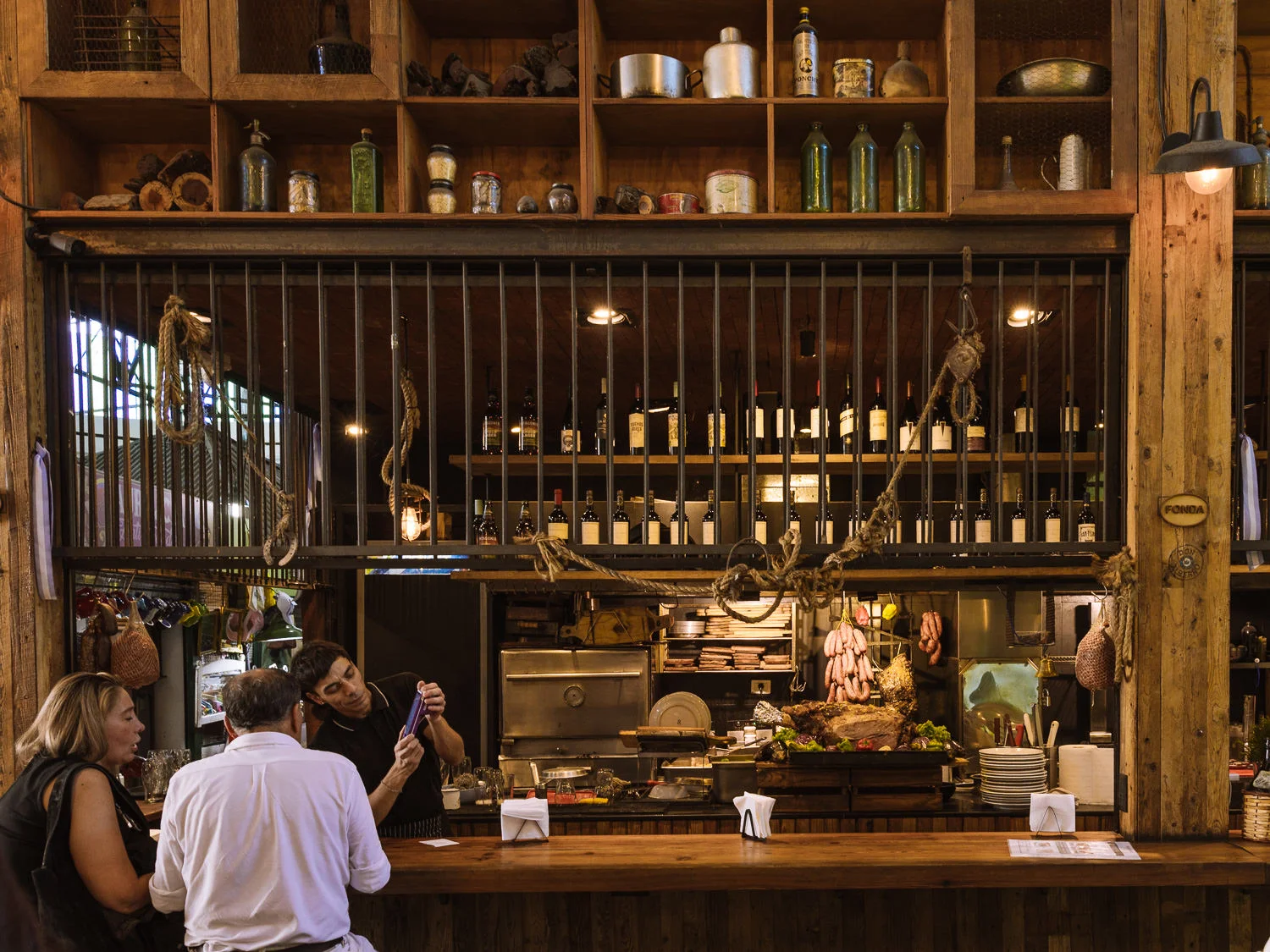 The rustic interior of a Buenos Aires parrilla, with hanging sausages, cuts of meat on display, wine bottles lining the shelves, and diners seated at a wooden bar counter