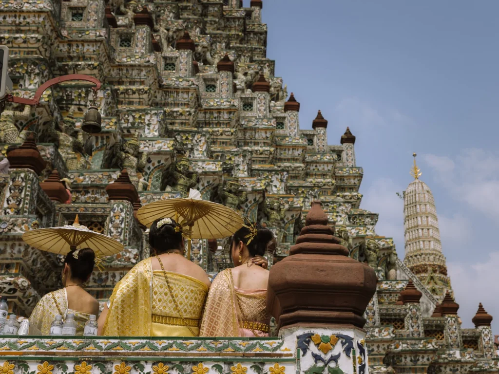 Three women in traditional Thai dress holding gold parasols stand before the ornate porcelain-encrusted prang of Wat Arun, Bangkok.