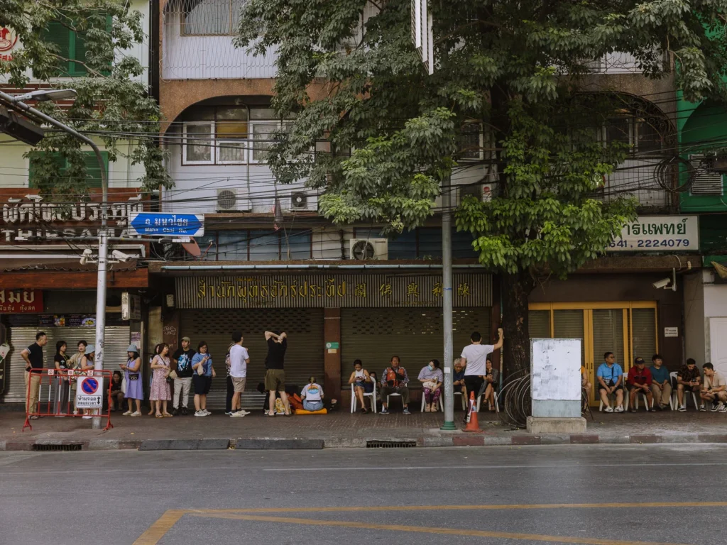 A crowd of people wait on the pavement outside a row of shophouses on Maha Chai Road, Bangkok.