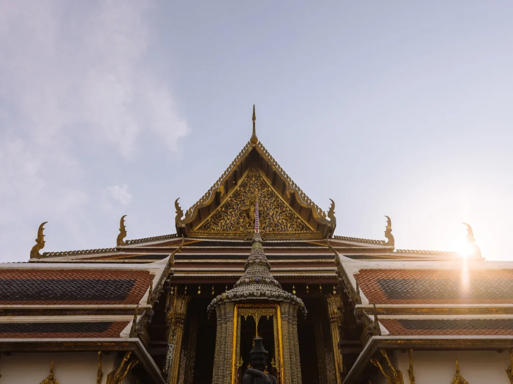 Looking up at the ornate gilded gable of a temple building within the Grand Palace complex, Bangkok, with sunlight flaring behind the roofline.