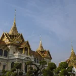 The Chakri Maha Prasat throne hall at the Grand Palace, Bangkok, its European neoclassical facade topped with ornate Thai spires against a blue sky.