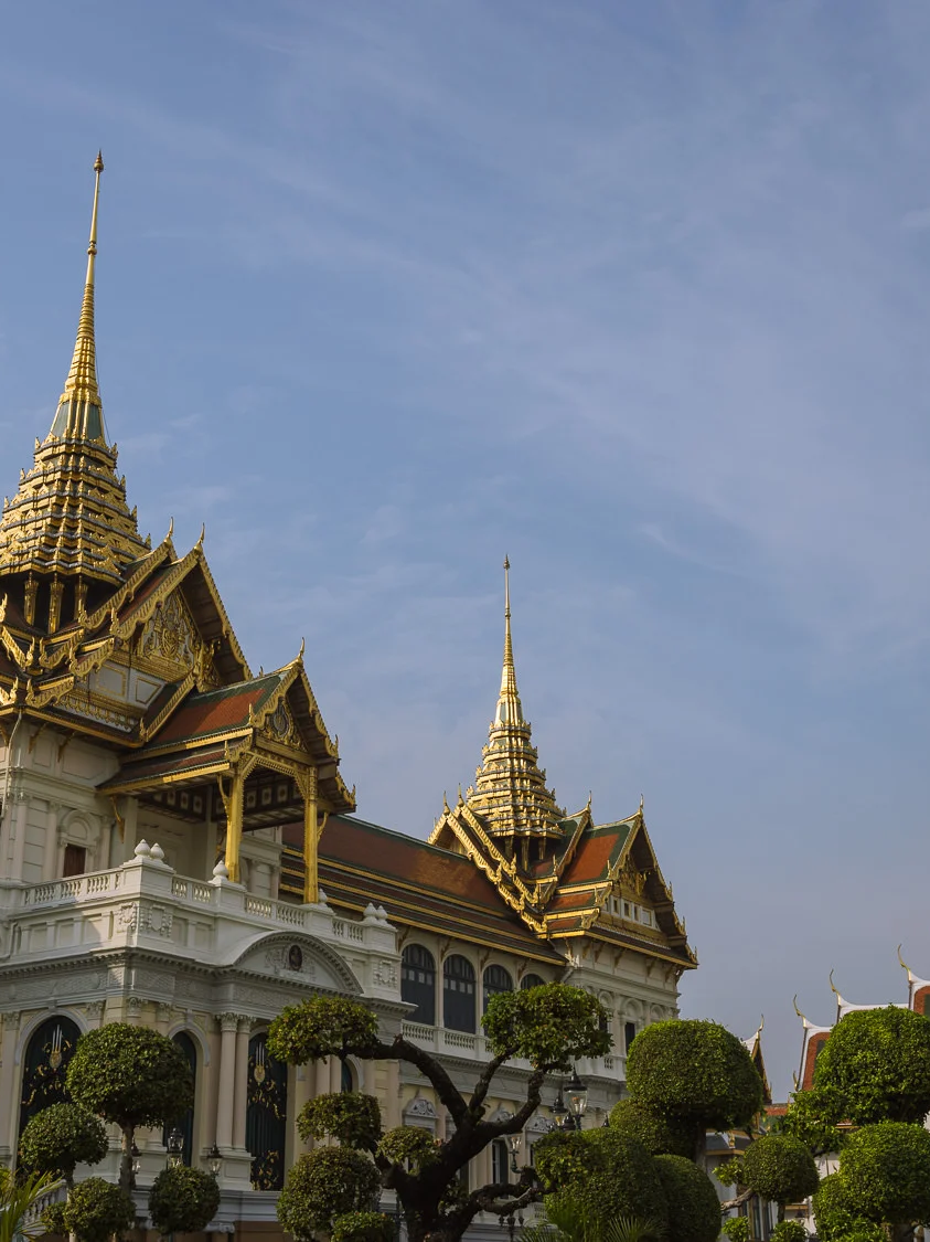 The Chakri Maha Prasat throne hall at the Grand Palace, Bangkok, its European neoclassical facade topped with ornate Thai spires against a blue sky.