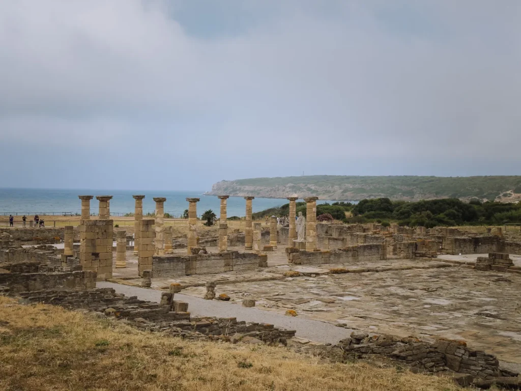 The ancient Roman ruins of Baelo Claudia near Tarifa, showing stone columns, wall foundations, and a white marble statue, with the Atlantic Ocean and green headland visible in the background