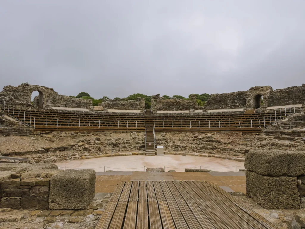 The semicircular ruins of the Roman theatre at Baelo Claudia near Tarifa, with restored wooden seating tiers set into the ancient stone cavea and a sandy arena floor below under an overcast sky