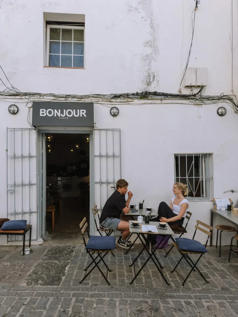 A couple sitting at a folding table with coffee cups outside Bonjour Tarifa café, a minimalist white-walled venue with a grey sign, industrial wall lights, and cobblestone pavement in the old town