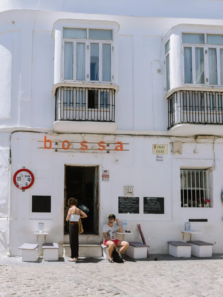 The white-walled exterior of Bossa bar-café on Calle Silos in Tarifa, with an orange logo sign, chalkboard menus, and two people relaxing outside on a bright sunny day