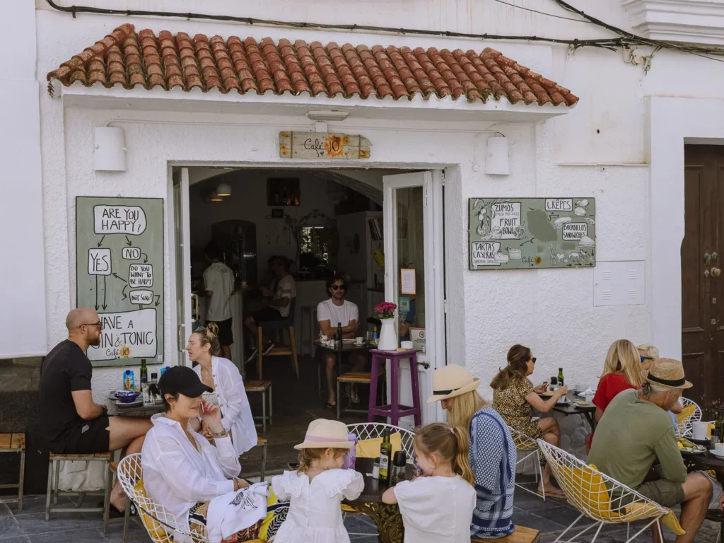 A busy outdoor terrace at Café 10 in Tarifa's old town, with guests dining at wire chairs under a terracotta-tiled awning, a chalkboard menu reading "Are You Happy? Have a Gin & Tonic" on the left and a menu board on the right