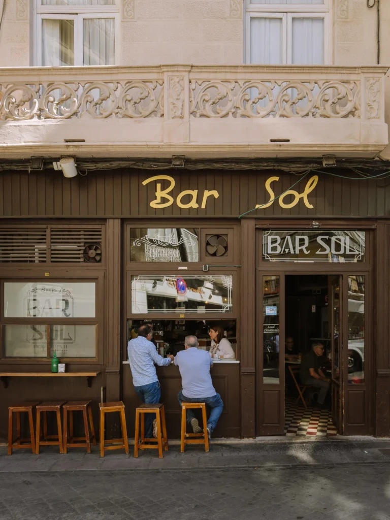 Two men standing at the outdoor counter of Bar Sol, a traditional tapas bar in Cartagena, Spain, with dark wood panelling, gold lettering, ornate ironwork above, wooden bar stools outside, and a glimpse of the chequered tile floor inside