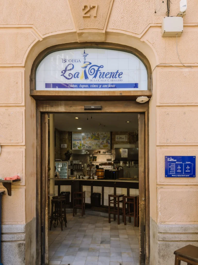 The arched doorway entrance of Bodega La Fuente de la Calle Caballero in Cartagena, Spain, with a tiled sign advertising cañas, tapas, vinos y anchoas, wooden bar stools visible inside, and colourful illustrated tiles decorating the back wall