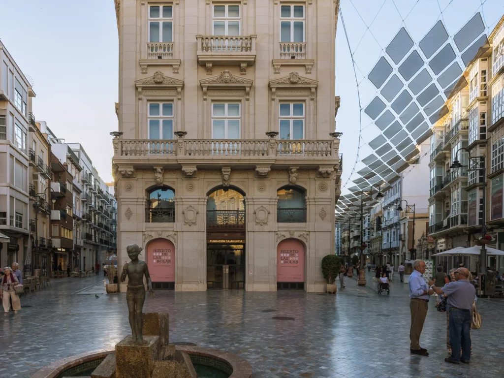 The pedestrianised Calle Mayor in Cartagena, Spain, at dusk, with the ornate Eclectic-style Fundación CajaMurcia building at its head, a bronze fountain statue in the foreground, locals gathered in conversation, and the city's distinctive solar shade canopy stretching overhead against a blue evening sky
