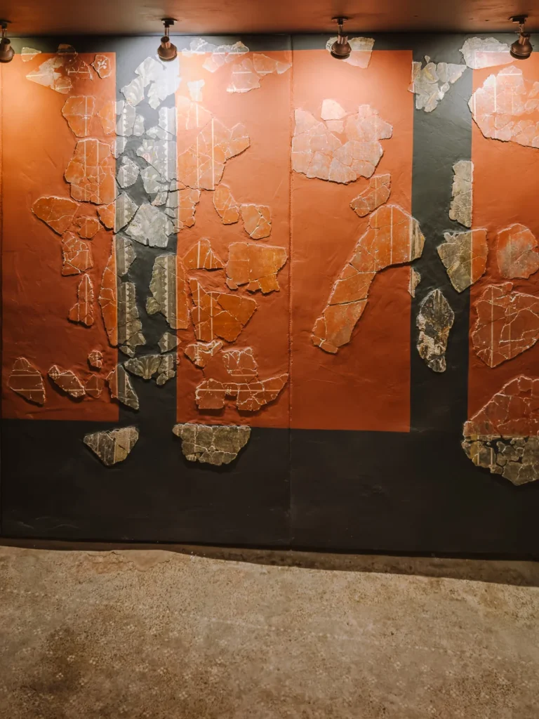 Fragments of original Roman fresco plasterwork mounted on a dark display wall at the Casa de la Fortuna in Cartagena, Spain, showing terracotta red and grey painted sections with delicate botanical motifs and geometric border designs