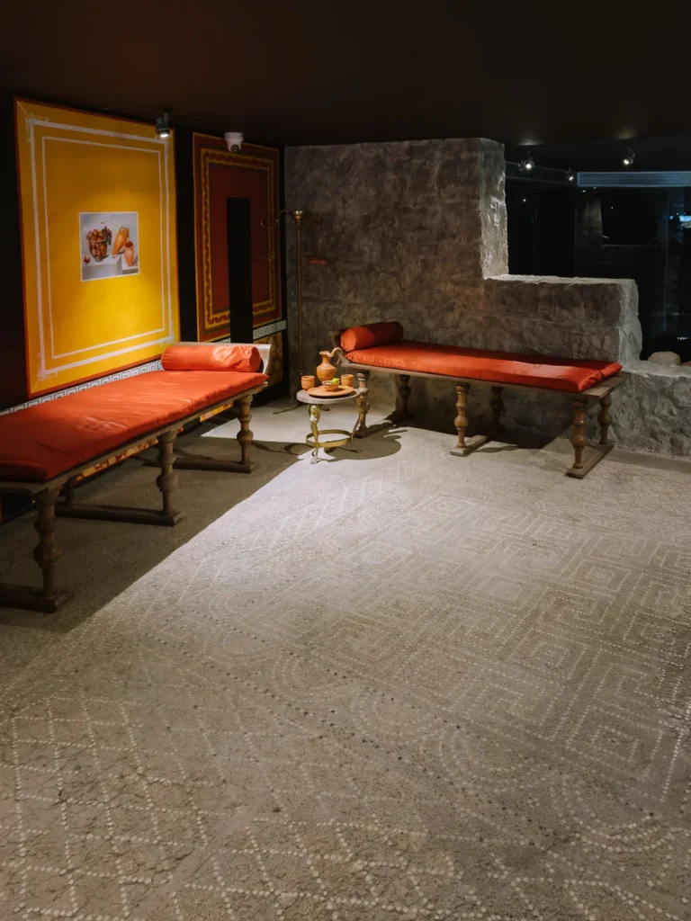 A reconstructed Roman triclinium (dining room) at the Casa de la Fortuna in Cartagena, Spain, with two red-cushioned day beds, a small bronze table set with clay vessels, vivid yellow and red fresco-style wall panels, and an original geometric white mosaic floor beneath