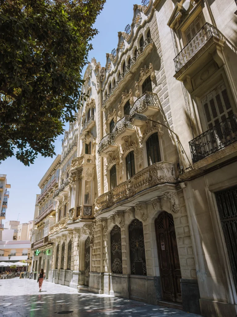The extravagant Modernista facade of Casa Maestre on Calle Mayor in Cartagena, Spain, viewed from street level, with densely carved white stone balconies, ornate floral ironwork, scalloped gable detailing, and a lone pedestrian walking below, framed by a ficus tree to the left