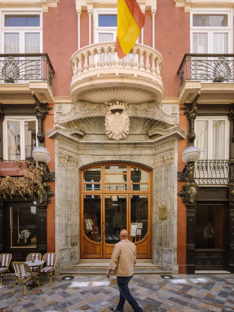 The ornate entrance of the Casino de Cartagena, Spain — a Modernista building featuring a carved stone doorway, decorative ironwork, and a balcony draped with the Spanish flag