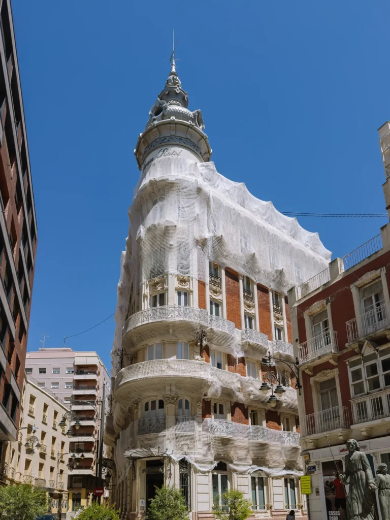 The Gran Hotel de Cartagena viewed from street level, its ornate Modernista corner tower and brick facade draped in white protective netting to prevent debris falling, with a bronze sculpture group visible to the right and a deep blue sky above