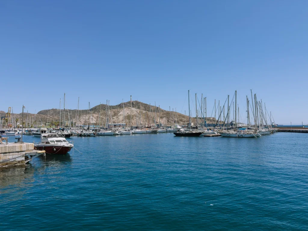 The natural deep-water harbour of Cartagena, Spain, with dozens of sailboats and yachts moored in the marina, their masts reflected in the calm turquoise water, and a bare rocky hill rising behind the moorings under a cloudless blue sky