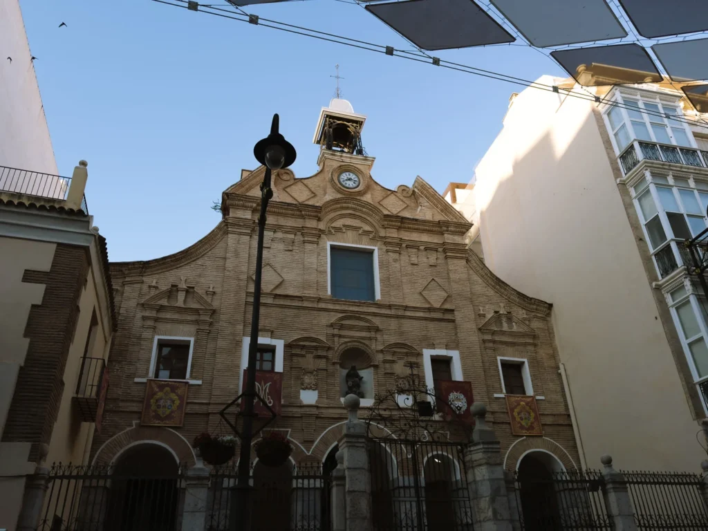The Baroque facade of the Iglesia de Santa María de Gracia in Cartagena, Spain, featuring a stepped gable, ornate stone detailing, a clock face, an open bell tower, and decorative religious banners hanging at the entrance, with a modern shade canopy visible overhead