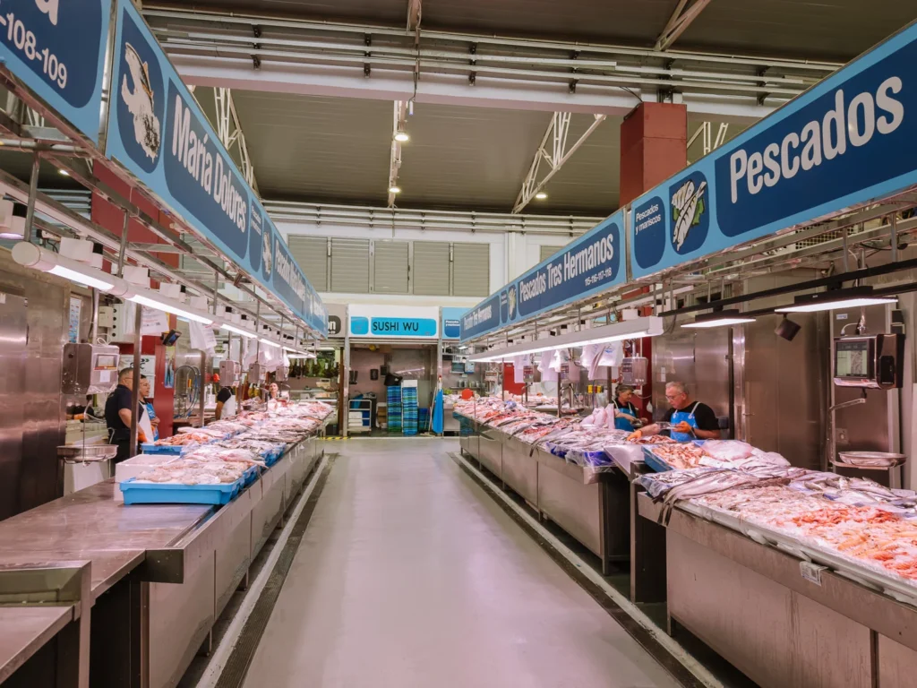 The fresh fish and seafood aisle inside the Mercado de Santa Florentina in Cartagena, Spain, with vendors preparing prawns, whole fish, and shellfish on stainless steel counters beneath blue "Pescados" signs, including stalls for María Dolores, Pescados Tres Hermanos, and a Sushi Wu counter at the far end
