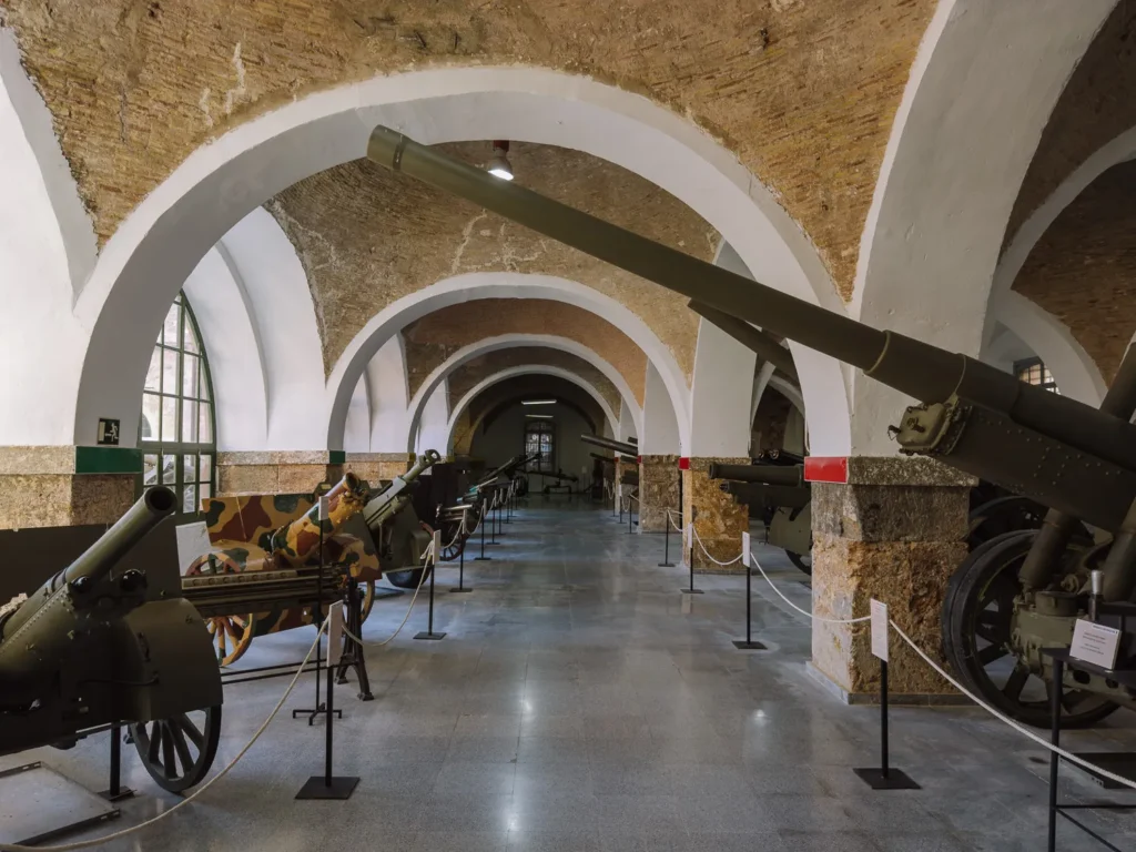A vaulted gallery inside the Museo Histórico Militar de Cartagena, Spain, displaying a row of historic artillery pieces and field guns beneath white-plastered brick arches in the 18th-century Real Parque y Maestranza de Artillería building