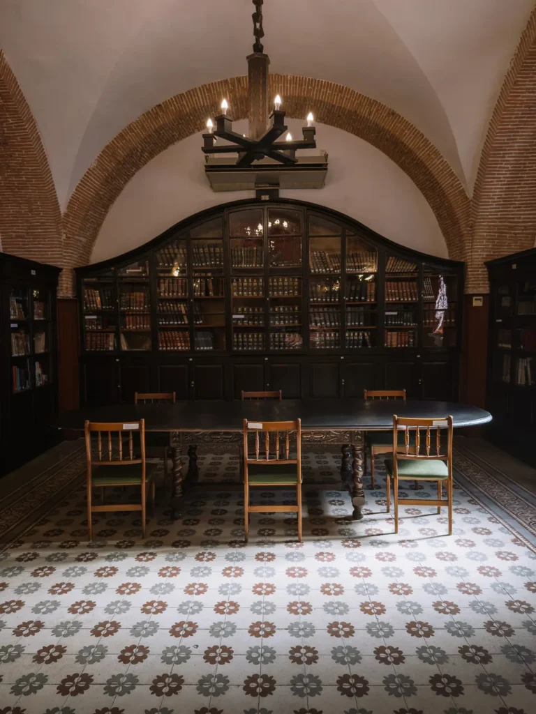 A dimly lit historic library room inside the Museo Histórico Militar de Cartagena, Spain, featuring floor-to-ceiling dark wood bookcases filled with old volumes, a large oval table with wooden chairs, a patterned tile floor, and a brick vaulted ceiling with a candelabra chandelier