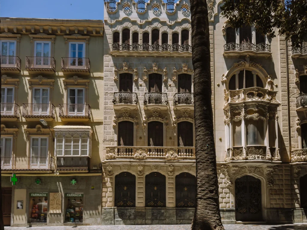 A row of ornate Modernista building facades on a sun-dappled street in Cartagena, Spain, including the elaborately decorated Casa Maestre and Casa Pedreño, with intricate stone carvings, wrought iron balconies, and arched ground-floor shopfronts