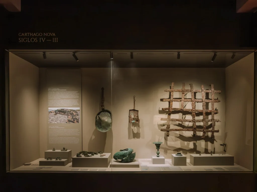 A dimly lit museum display case labelled "Carthago Nova — Siglos IV–III" inside the Roman Forum at Molinete Hill Cartagena, Spain, featuring ancient Carthaginian bronze artefacts including a helmet, tools, a decorative chalice, and a large iron grate structure