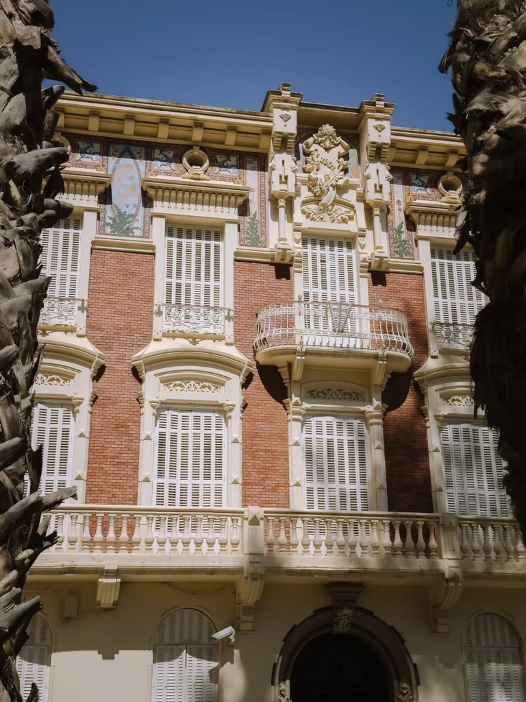 The ornate Modernista facade of the Palacio de Aguirre in Cartagena, Spain, framed by palm trees, featuring red brick with elaborate white plasterwork detailing, decorative painted tile panels, arched louvred shutters, wrought iron balconies, and a central carved crest above the entrance arch