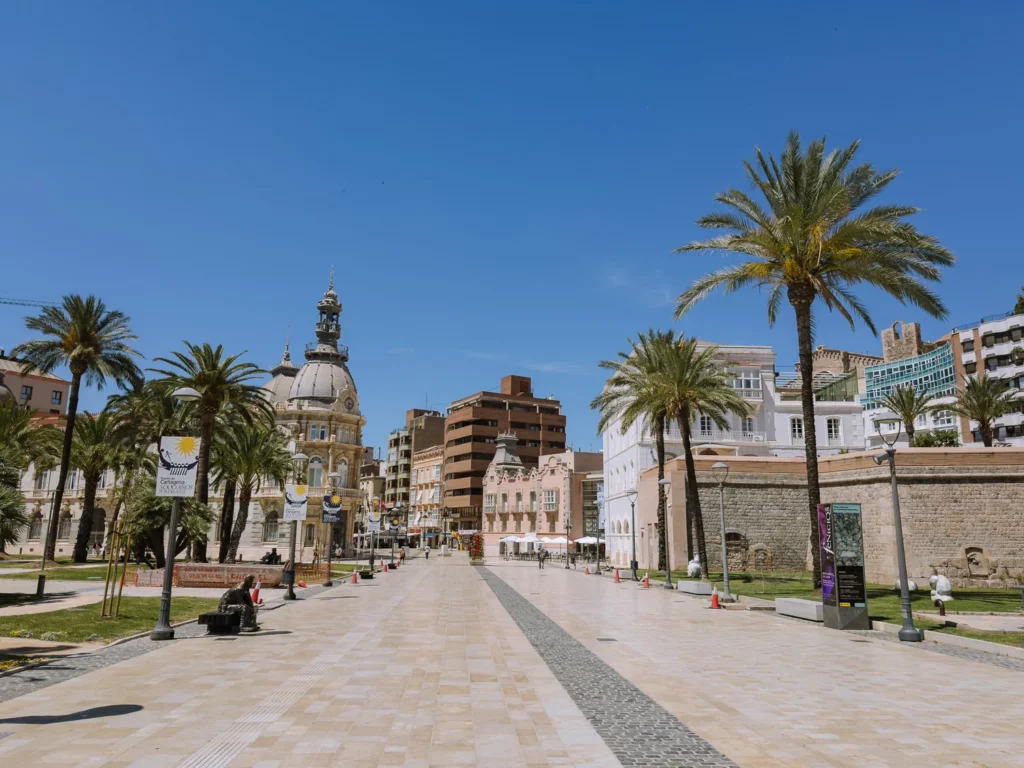 The broad, marble-paved Plaza del Ayuntamiento in Cartagena, Spain, lined with tall palm trees under a deep blue sky, with the domed Palacio Consistorial visible to the left, ancient city walls to the right, and a lone bronze sculpture seated on a bench in the middle ground