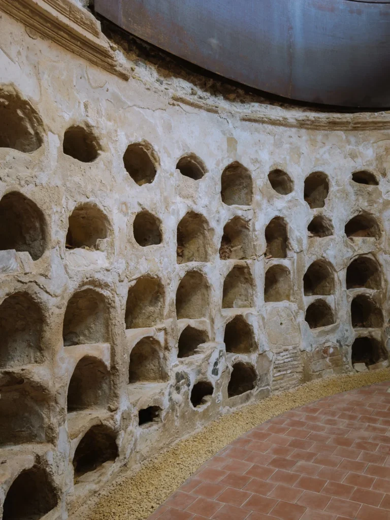 The curved interior wall of the Hermitage of San José in Cartagena, Spain, lined with rows of arched stone niches, preserved beneath a modern steel canopy with terracotta floor tiles in the foreground