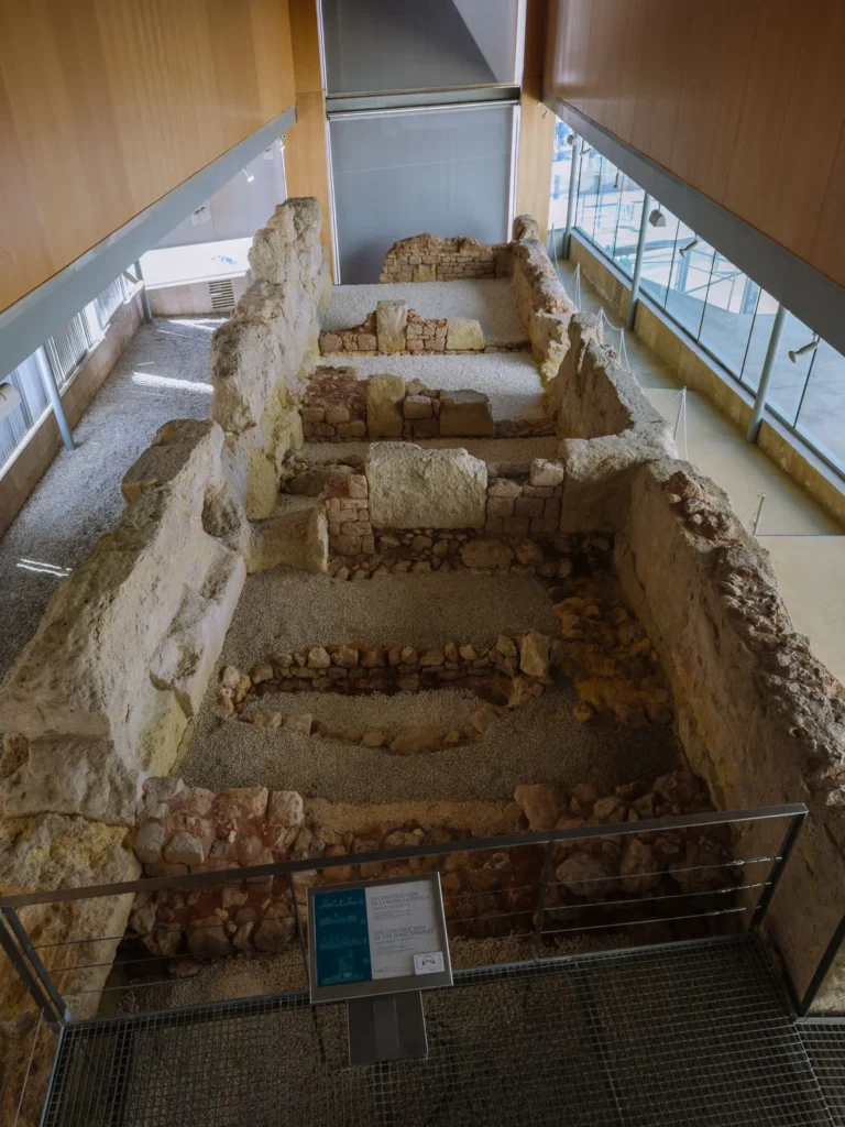 The excavated remains of the ancient Punic Wall of Cartagena, Spain, viewed from an elevated walkway inside the Museo del Teatro Romano, showing the original Carthaginian stone foundations preserved beneath the modern building with interpretation panels alongside
