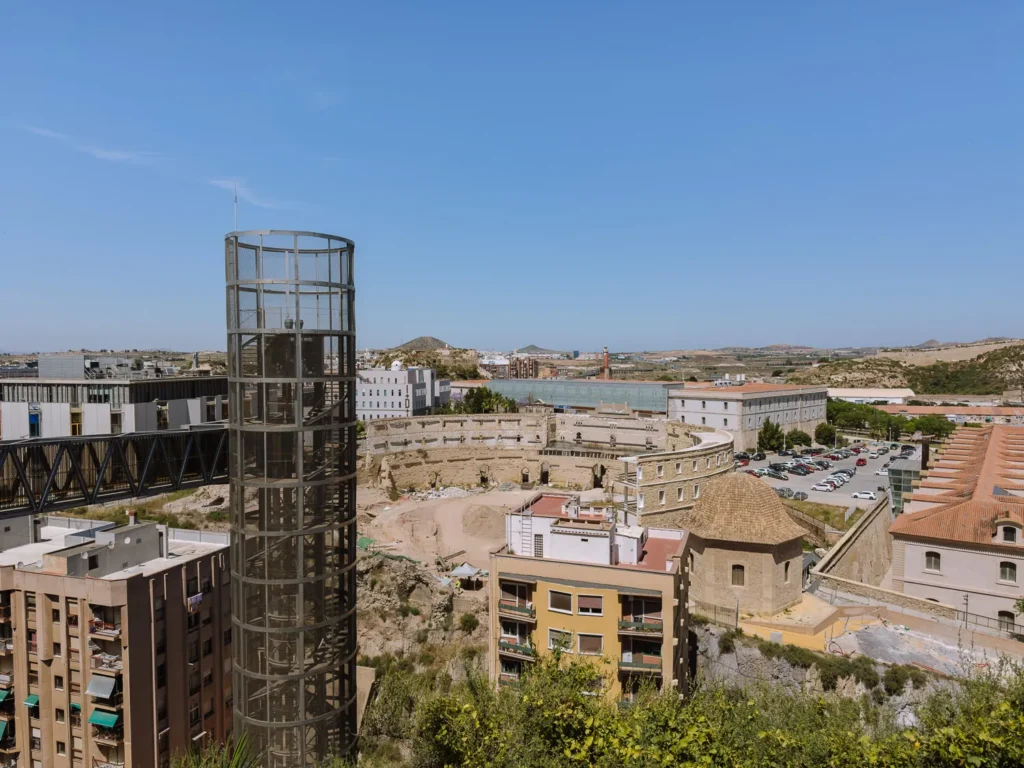 The Roman Amphitheatre excavation site in Cartagena, Spain, viewed from above, with the modern glass panoramic lift tower in the foreground, the elliptical ruins of the ancient arena and 19th-century bullring in the middle ground, and the city stretching to the hills beyond