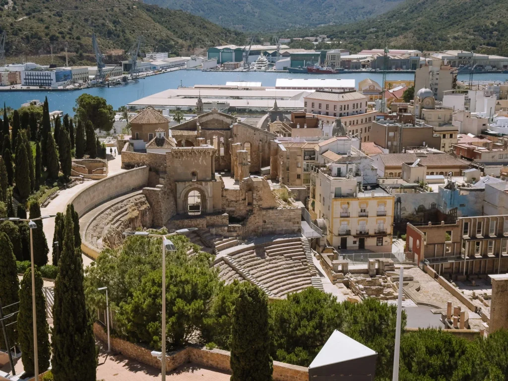 An aerial view of the Roman Theatre of Cartagena from the Castillo de la Concepción, showing the sweeping semicircular stone seating, the ruined facade of the Cathedral of Santa María la Vieja behind it, and the city's deep-water naval harbour stretching into the distance beneath the surrounding green hills