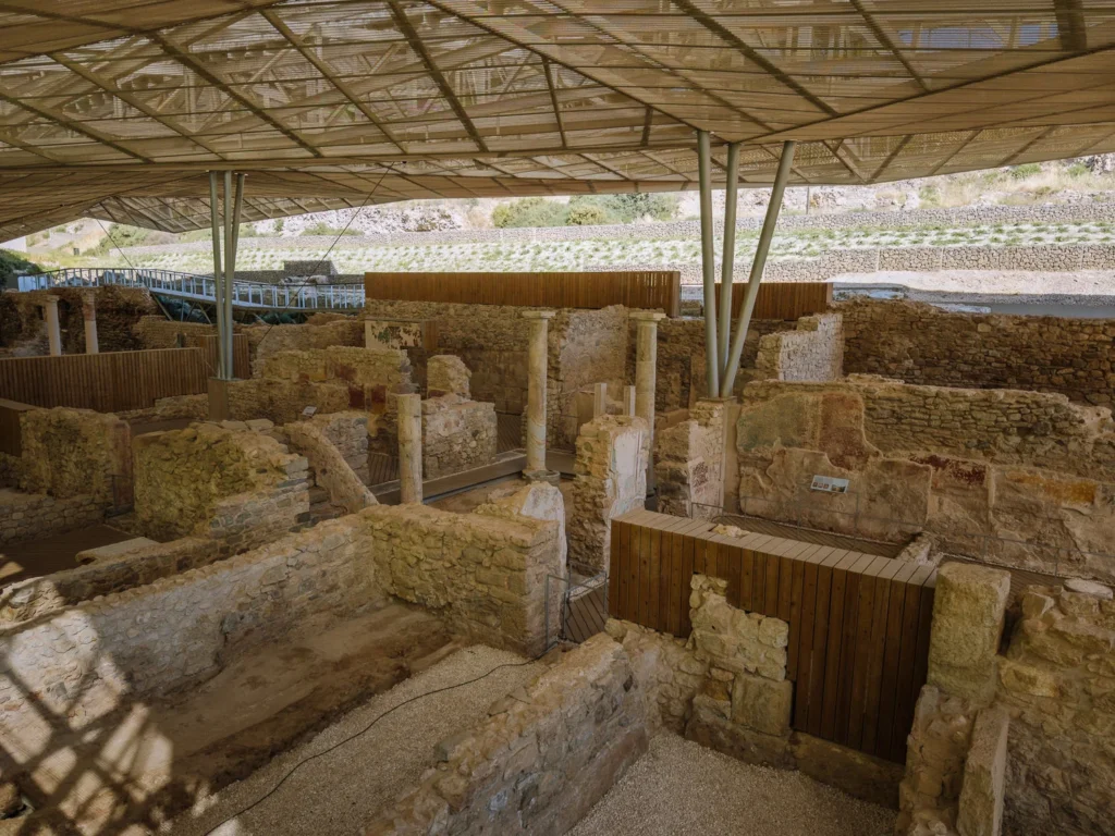 The excavated ruins of a Roman villa near Cartagena, Spain, protected by a large modern canopy structure, with stone walls, column bases, and wooden walkways winding between the exposed ancient foundations, set against a dry hillside landscape