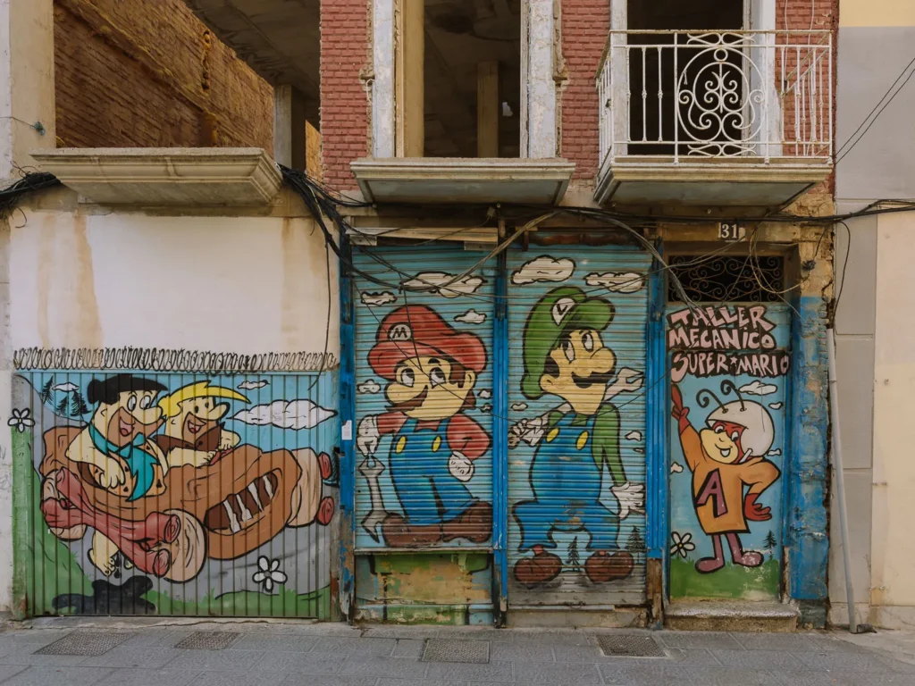 Colourful street art painted on roller shutters in Cartagena, Spain, featuring cartoon characters including Mario and Luigi from Super Mario Bros and Fred and Barney from The Flintstones, labelled "Taller Mecánico Super Mario"
