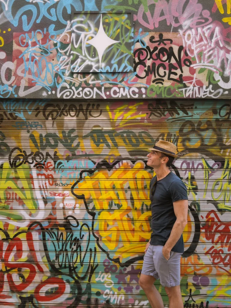 A man in a straw hat and navy t-shirt stands in front of a colourful graffiti-covered roller shutter on a street in Cartagena, Spain