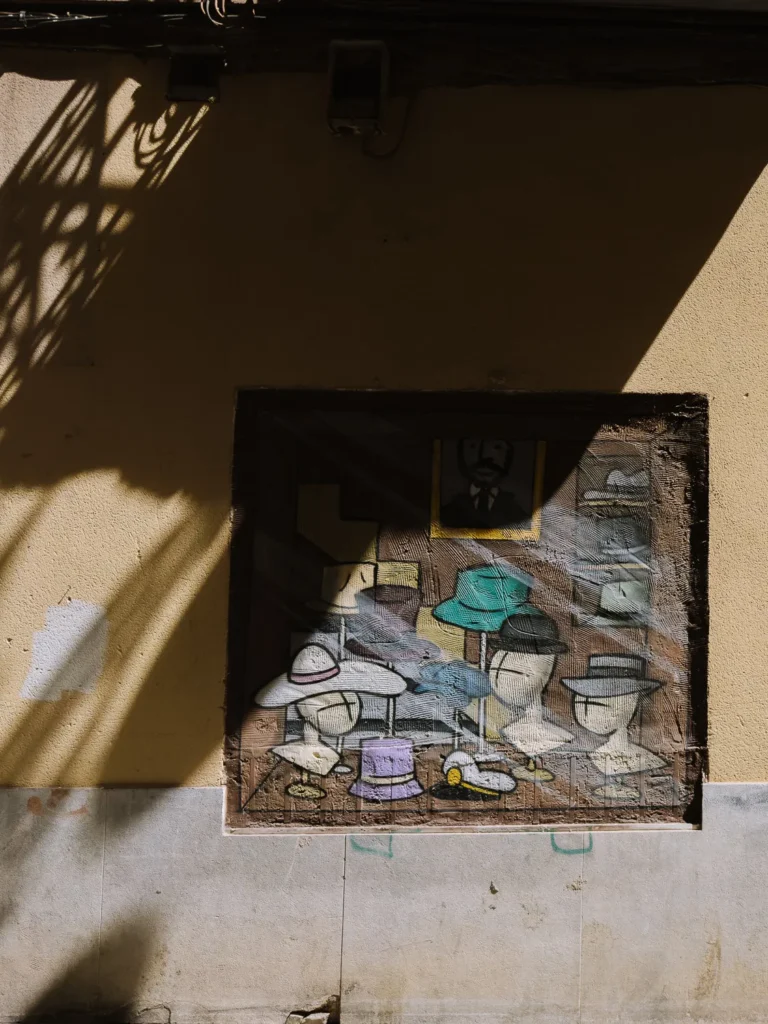 A detailed street art mural painted into a recessed wall niche in Cartagena, Spain, depicting a hat shop display with various styles of hats on mannequin heads, bathed in dappled sunlight